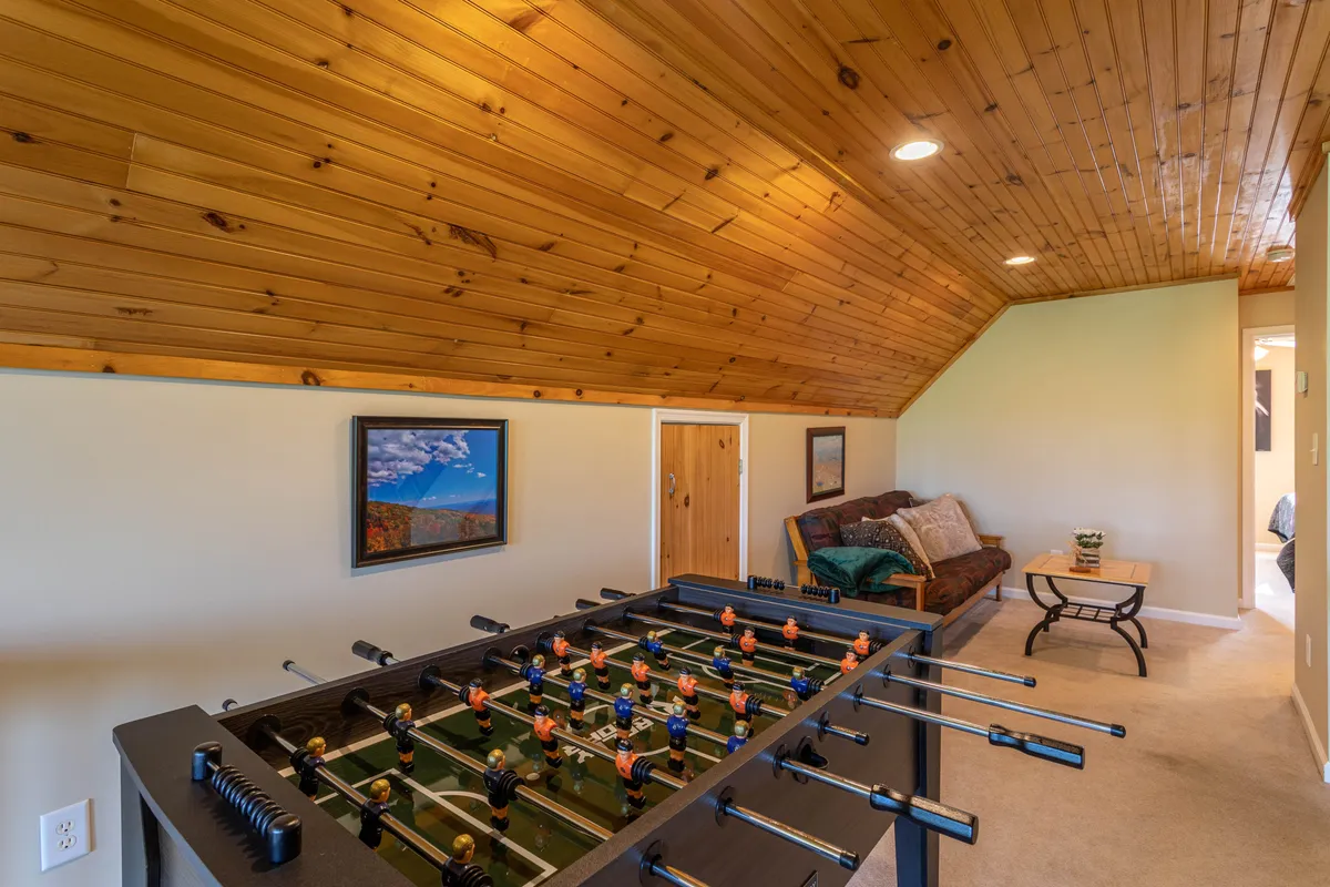 The loft is depicted from a slightly different angle, spotlighting the foosball table, a comfortable couch, wood-paneled ceiling, and framed artwork. The space feels playful yet welcoming, ideal for both active games and lazy lounging.