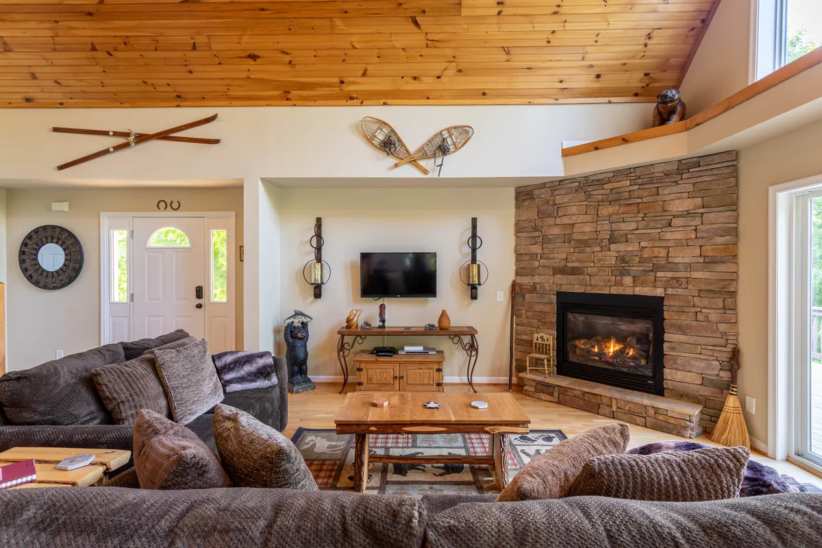 A warmly lit living area featuring a striking stone fireplace, exposed wooden beams, large windows offering glimpses of the outdoors, and plush seating arranged for relaxation. Decorative artifacts add a cozy, romantic ambiance to the room.