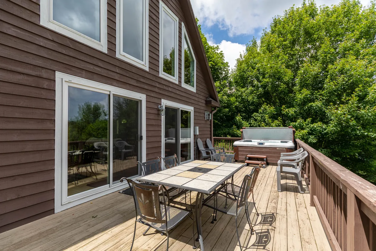 This photo shows a well-appointed deck with a dining table, hot tub, and views of greenery and large windows leading back into the home. The space is set up for outdoor dining, relaxation, and social gatherings, all in close proximity to nature.