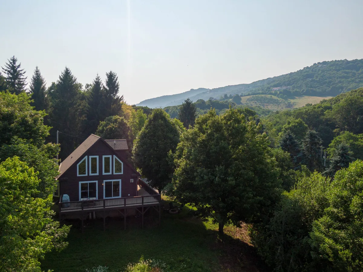 The image showcases a charming wooden cabin nestled among lush trees, with a spacious deck overlooking rolling mountains in the distance. The peaceful outdoor setting is ideal for relaxing, taking in sunrise views, or gathering under the stars.