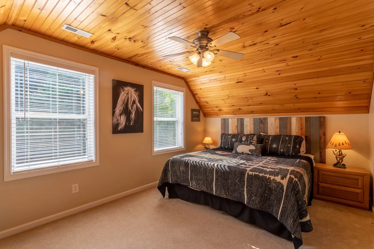An inviting bedroom with a rustic wooden ceiling, a king bed adorned with patterned bedding, decorative equestrian artwork above the headboard, and soft lighting from a table lamp. Natural light filters in, reinforcing the peaceful retreat atmosphere.