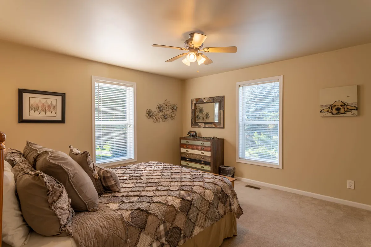 A warmly lit bedroom with a large bed adorned in patterned bedding, a wooden dresser, framed wall art, and windows allowing natural light to spill in. The decor is inviting, with touches that evoke both comfort and a connection to the surrounding landscape.