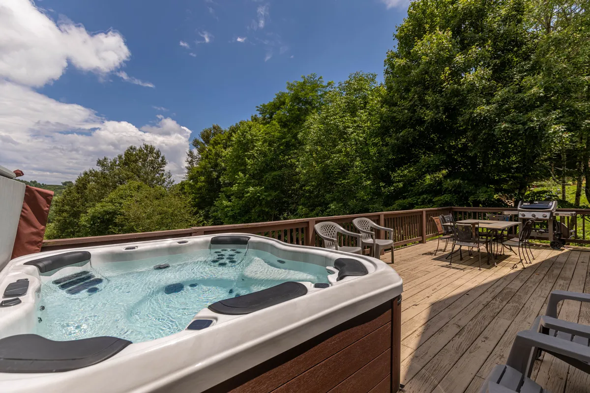 This image captures the wraparound deck featuring an inviting hot tub, comfortable outdoor seating, and lush greenery as the backdrop. The scene conveys a tranquil, social setting with the hot tub perfectly positioned for sunset views, and the space is ideal for entertaining or soaking in the scenery.
