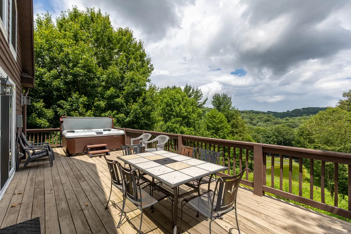 The image displays a broad view of the wooden deck, complete with a large dining table, hot tub, and surrounding greenery. This inviting scene captures morning coffee moments, alfresco dining, and evening soaks with beautiful natural surroundings.