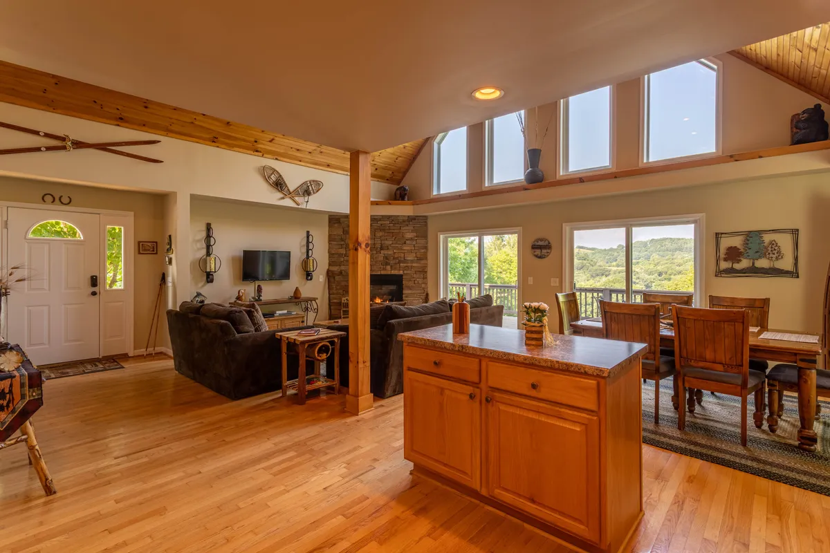 A spacious, open-concept living area featuring a central kitchen island with wooden cabinetry, a comfortable seating area arranged around a stone fireplace, and expansive windows that frame panoramic mountain views. The space is bathed in natural light, making it ideal for gathering with family and friends for meals, conversation, or relaxing after a day outdoors.