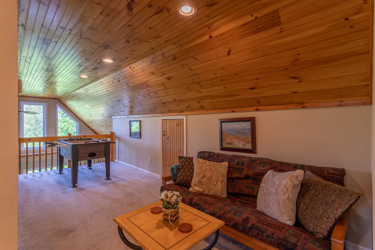 This image captures a bright, inviting loft space featuring a comfortable sofa, a foosball table at the center, and decorative touches like artwork and a flower-adorned table. Large windows in the background suggest a connection to scenic views, while the layout balances playfulness and relaxation.