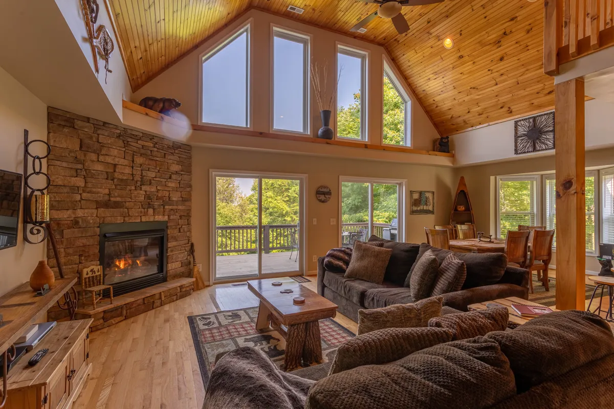 A welcoming living room with a large stone fireplace as the focal point, surrounded by comfortable seating, wooden beams overhead, and expansive windows that let in ample natural light. The view outside hints at mountain scenery, reinforcing the lodge’s cozy yet scenic ambiance.